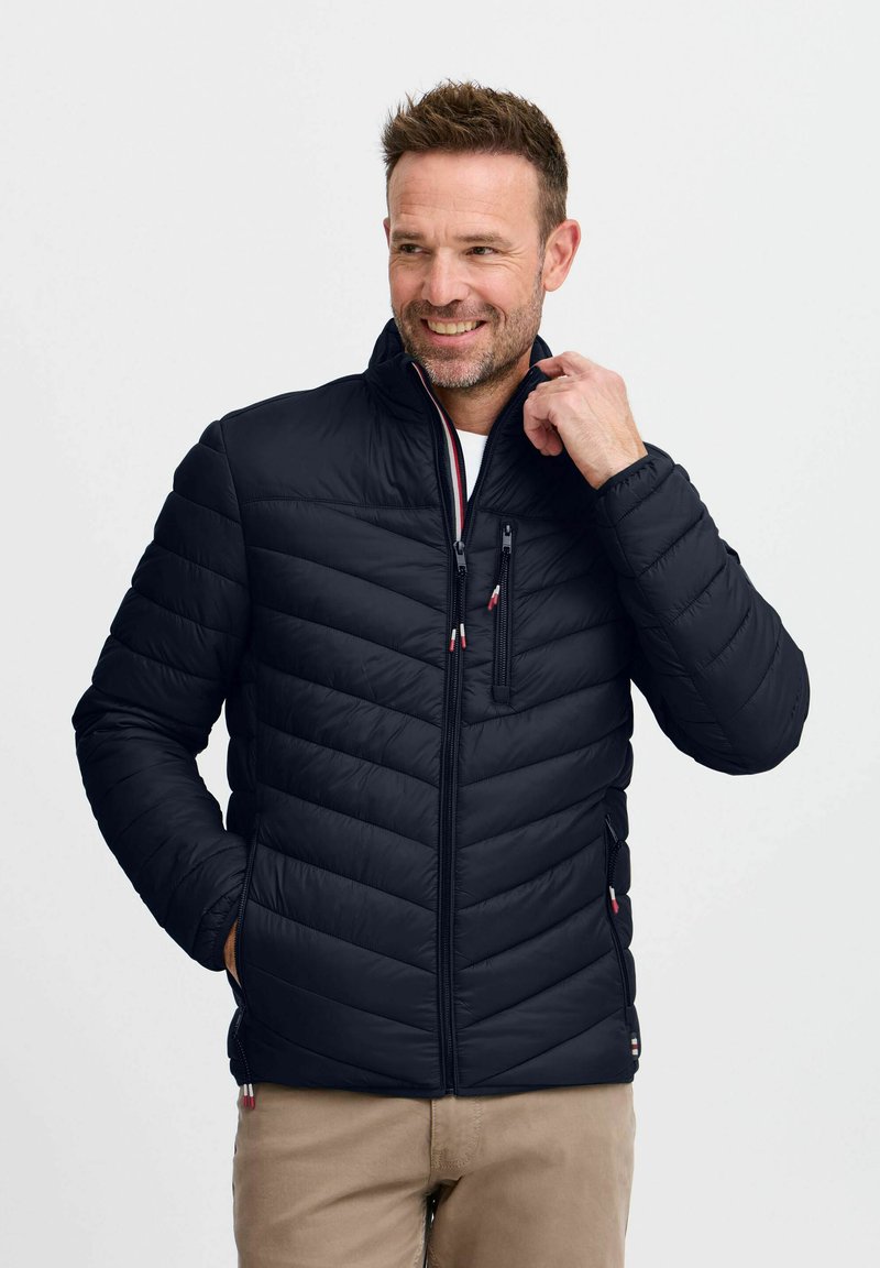 Chaqueta acolchada negra con un alto cuello, que presenta un diseño de acolchado horizontal, bolsillo en el pecho con cremallera y acentos en contraste en las cremalleras.