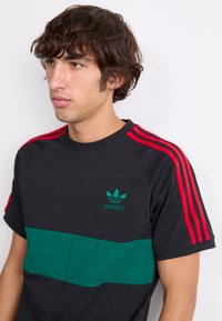Juodas marškinėliai su žalia horizontalia juostele ir mažuoju žaliu Adidas logo. Rankovėse yra raudonos juostos. Minkšta medvilnė. Kasdienis švarkas.