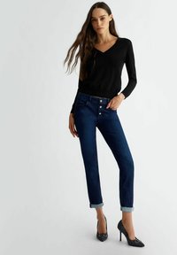 Jeans in denim blu scuro con gamba affusolata, dotati di zip con bottoni e polsini arrotolati. Abbinati a un maglione nero a maniche lunghe con scollo a V e scarpe con tacco.