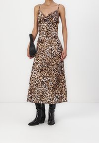 Robe longue en satin à imprimé léopard avec col bénitier et fines bretelles, associée à des bottes noires hautes jusqu'aux genoux et un petit sac à main noir. Texture lisse.