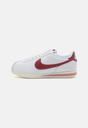 Baskets blanches Nike Cortez avec logo Swoosh rouge et accents rouges, semelle plate et lacets blancs, vues de profil sur fond blanc.
