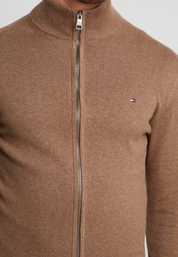 Brun strikket zip-up sweater med et lille rektangulært rød, hvidt og blåt logo på venstre bryst, båret af en person.