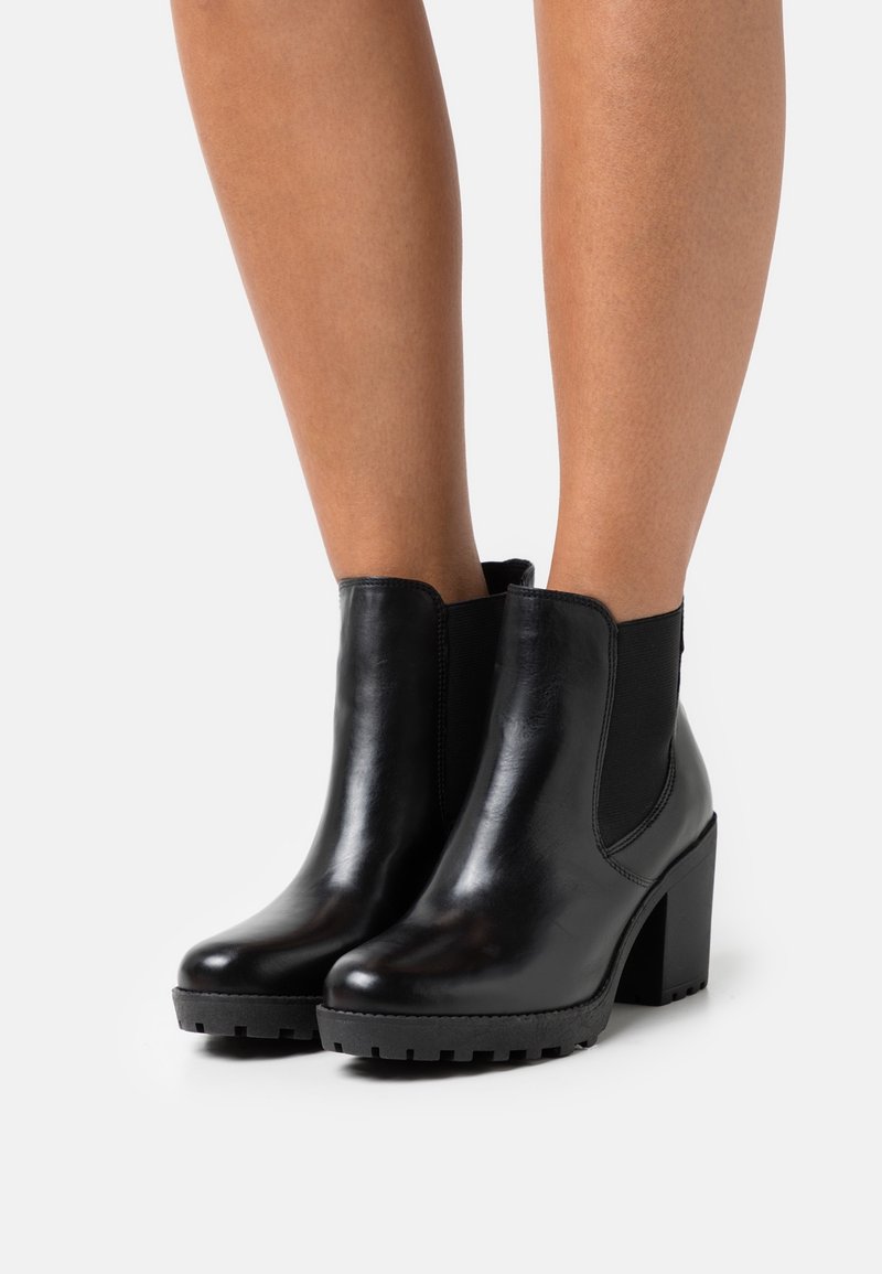 Bottes courtes en cuir noir avec talons hauts épais et panneaux latéraux élastiques, portées sur des jambes nues sur un fond uni.
