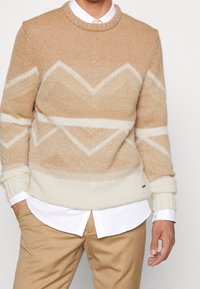 Pull beige en maille avec un motif chevron dans des teintes plus claires, doté de poignets et d'un ourlet côtelés, associé à une chemise blanche à col.