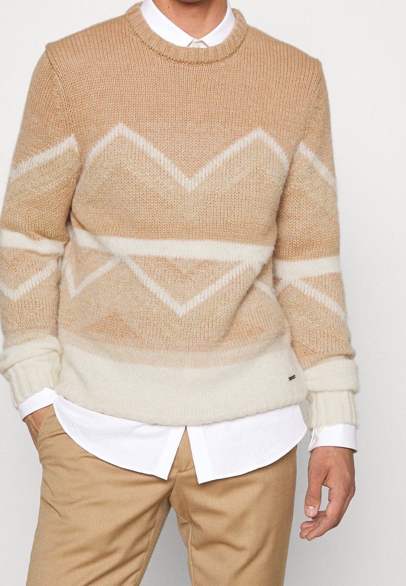 Pull beige en maille avec un motif chevron dans des teintes plus claires, doté de poignets et d'un ourlet côtelés, associé à une chemise blanche à col.