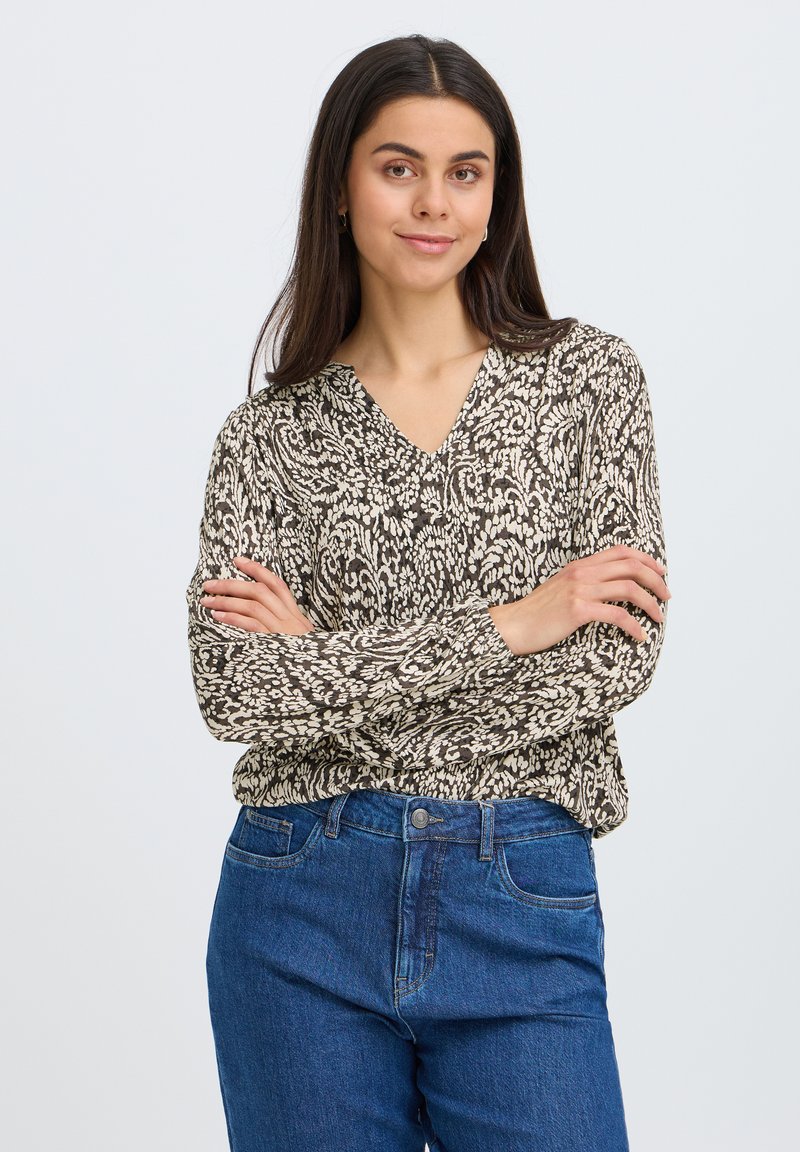 Femme aux longs cheveux foncés, bras croisés, portant un chemisier à motifs et un jean bleu, souriant devant un fond uni.