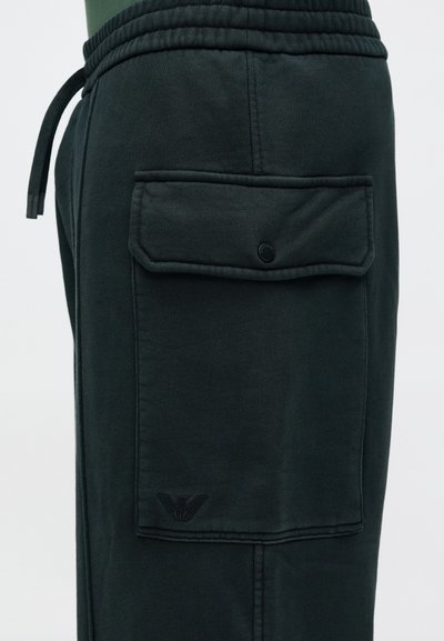 Tmavozelené tepláky z bavlnenej zmesi, so bočným vreckom na gombík a malým vyrazeným logom pri okraji nohavice.