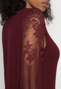 Bordeaux top met een getailleerd lichaam en doorzichtige kanten mouwen met bloemenpatronen en verticale strepen. Soepele textuur en elegant ontwerp.