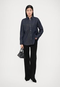 Modrá tmavomodrá rifľová košeľa na gombíky s dvoma náprsnými vreckami, v kombinácii s čiernymi nohavicami s vysokým pásom. V ruke čierna štruktúrovaná kabelka.