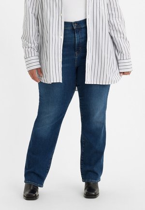 Personne portant un jean droit bleu foncé, des bottes noires, un haut blanc et une chemise blanche boutonnée avec de fines rayures noires.
