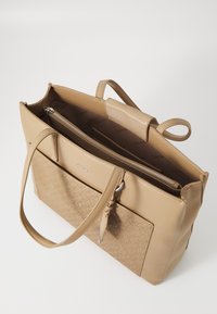 Sac cabas beige avec surfaces lisses et texturées. Comprend une poche zippée, des poignées supérieures, et un logo détaillé à l'avant. Intérieur spacieux.