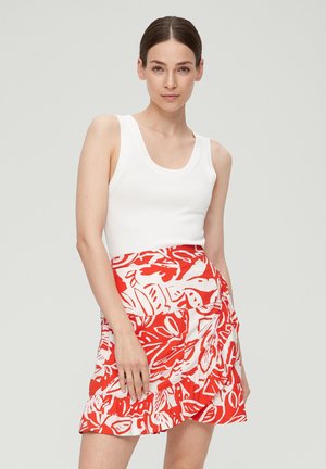 Vrouw met een lichte huid en donker haar in een witte mouwloze top en een rood-wit gestreepte rok, staande tegen een effen lichte achtergrond.