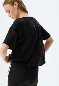 Musta urheilupaita lyhyillä hihoilla, jossa on verkkokangas selkäosa, pyöreä helma ja koristeelliset pistekoristeet kaula-aukossa.