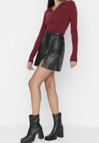 Haut à manches longues bordeaux associé à une mini-jupe en cuir noir avec une fermeture éclair sur le côté et une fente haute, complété par des bottines noires à talons.