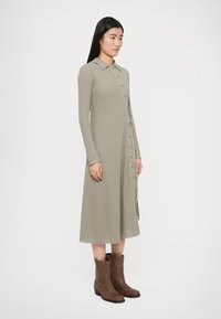 Khaki-ribbed kjole med krave, lange ærmer og knaplukning, kombineret med brune ankelstøvler. Glat tekstur og midt-lang længde.