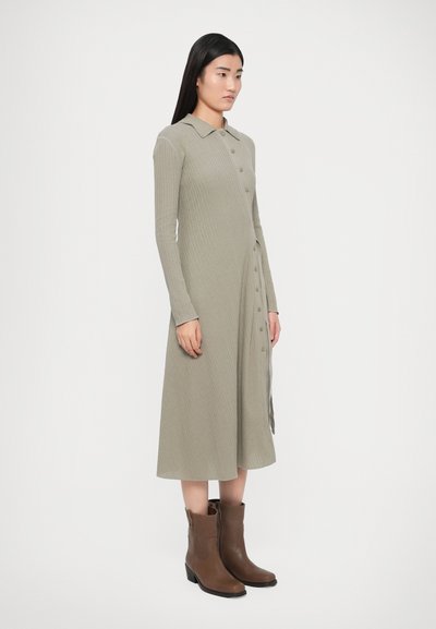 Robe côtelée kaki avec col, manches longues et fermeture par boutons, associée à des bottines marron qui montent jusqu'à la cheville. Texture lisse et longueur mi-mollet.