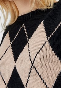 Suéter de punto con un patrón de diamante en negro y beige, con un cuello acanalado, que muestra una tela texturizada.