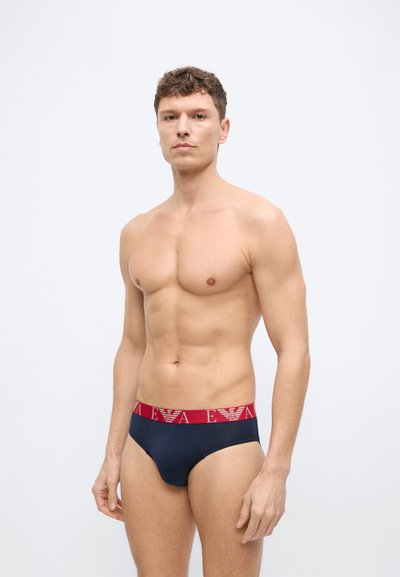 Άνδρας χωρίς πουκάμισο με κοντά καστανά μαλλιά, φοράει σκούρα μπλε εσώρουχα με κόκκινη μέση που έχει λευκό κείμενο και λογότυπα, στέκεται μπροστά σε απλό άσπρο φόντο.