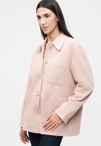 Γυναίκα που φοράει ανοιχτό ροζ μπουφάν με κουμπιά και τρεις μπροστινές τσέπες, στέκεται με το ένα χέρι να ακουμπά στην κάτω τσέπη.