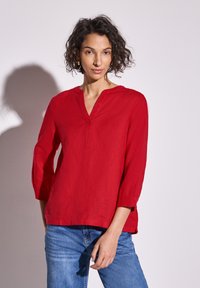 Blusa di lino rossa con scollatura a V e maniche a tre quarti, con fessure laterali per una maggiore libertà di movimento; tessuto morbido. Indossata con jeans blu.