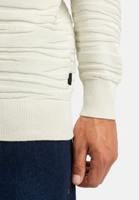 Maglione bianco testurizzato con righe orizzontali, polsini e orlo a costine. Etichetta visibile di lato. Indossato con jeans in denim scuro.