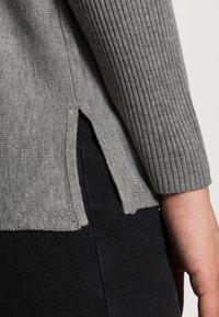 Pull gris en maille côtelée avec fentes latérales, manches longues et coupe décontractée, associé à un tissu noir texturé.