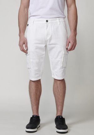 Pantalones cortos de carga blancos de algodón, con bolsillos laterales, cierre de botón y dobladillos enrollados. Llevados con zapatillas negras.