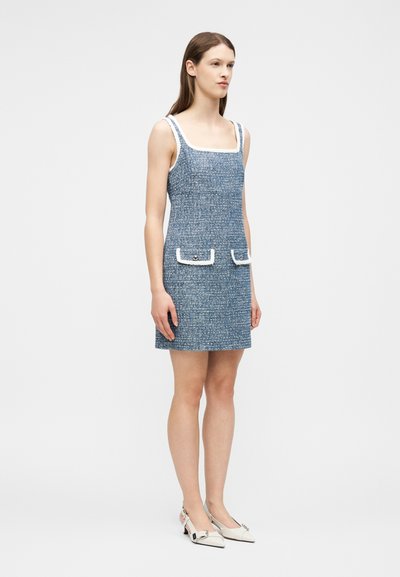 Femme portant une robe mini bleu tweed sans manches avec des bordures blanches et des poches avant, associée à des chaussures à bride arrière blanches et à petit talon.