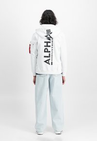 Sudadera blanca con gran impresión negra de "ALPHA INDUSTRIES" en la parte posterior, capucha ajustable y bolsillo en la manga. Combinada con vaqueros de pierna ancha de color azul claro.