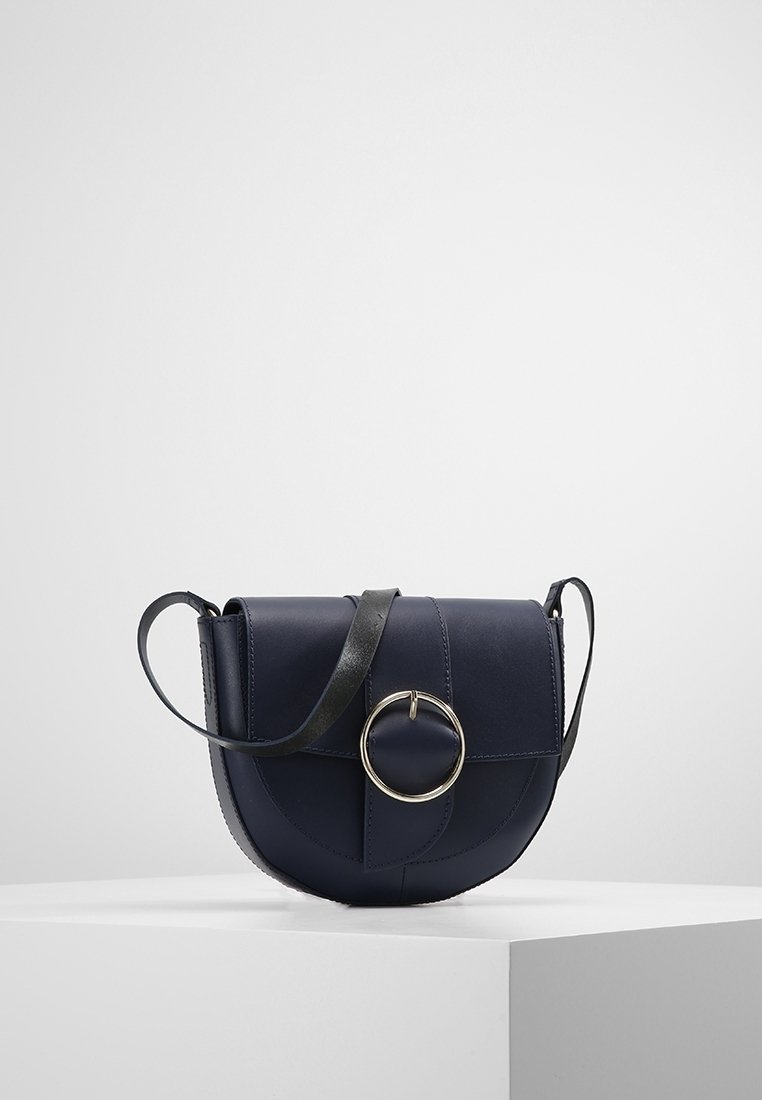 Borsa a tracolla in pelle blu navy con un accent metallici circolare, patta superiore piatta e una tracolla nera a contrasto. Base rotonda, texture liscia.