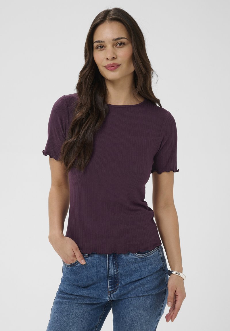 Femme aux longs cheveux foncés portant un haut violet à manches courtes avec des bords festonnés et un jean bleu, debout avec une main dans la poche.