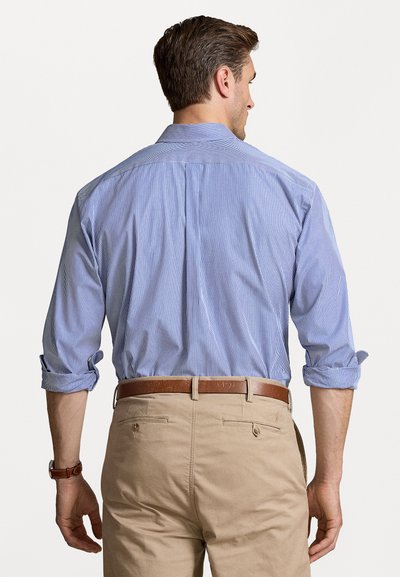 Chemise à rayures bleues avec des manches retroussées, associée à un pantalon beige et une ceinture marron, vue de dos. Texture de tissu lisse.