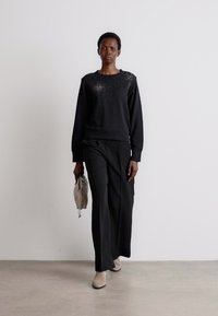 Melna džemperis ar studu detaļām, apvienots ar brīvām melnām biksēm. Modelis tur gaiši pelēku salocītu clutch somu. Beigē krāsas potīšu zābaki.