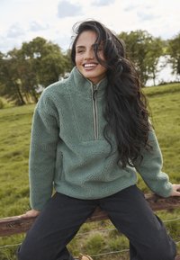 Pullover de forro polar verde con medio cierre frontal, textura acogedora y un bolsillo en el pecho. Combinado con pantalones oscuros, frente a un fondo natural.
