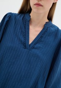 Kék, texturált blúz V-nyakkal és fodros díszítéssel a gallér mentén. Hosszú ujjak, ráncolt mandzsettával. Puha, könnyű anyag.