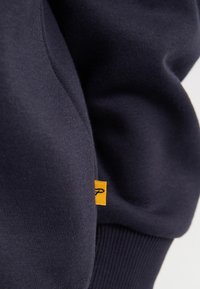 Felpa blu navy con una consistenza liscia, caratterizzata da un'etichetta gialla con un logo e polsini a coste. Il tessuto appare morbido e resistente.