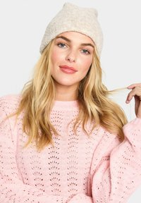 Cappello lavorato a maglia beige con la parte superiore arrotondata, abbinato a un maglione a maniche lunghe rosa pastello con un motivo di pizzo e maniche morbide.
