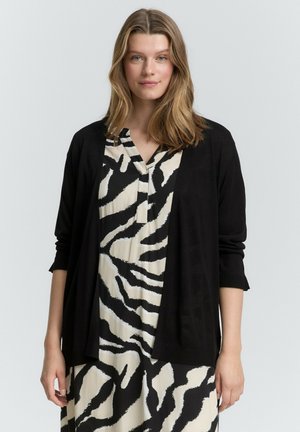 Cardigan noir en tissu doux, porté sur une robe blanche avec de boldes rayures zèbres noires. Présente une coupe décontractée et des manches trois-quarts.