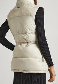 Beżowa kamizelka puchowa z kołnierzem stójkowym, pikowany wzór i boczne kieszenie. W połączeniu z czarną bluzką z długim rękawem z prążkowanego materiału.