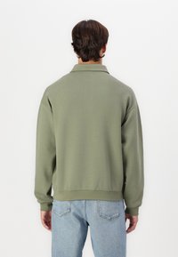 Sudadera de gran tamaño en color verde oliva con cuello, puños y dobladillo de canalé, combinada con jeans de color azul claro. Diseño simple y casual.