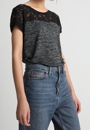 Mujer lleva una camiseta de manga corta negra y gris con detalles de encaje y vaqueros azules de talle alto sobre un fondo liso.