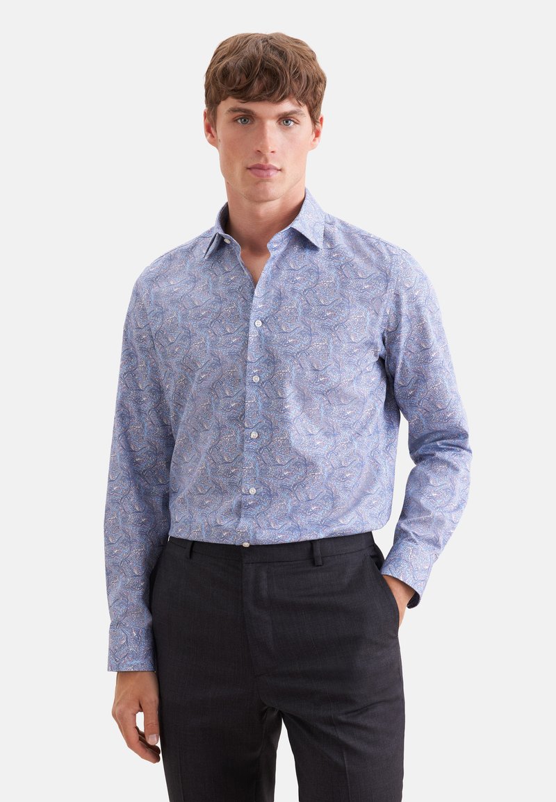 Jeune homme portant une chemise boutonnée à manches longues à motif paisley bleu et un pantalon noir, debout devant un fond clair uni.