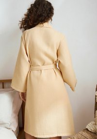 Beige, estruturált köntös, amely gofriszerű mintázattal rendelkezik, övvel a deréknál és széles ujjakkal. A ruha a vádli közepéig ér, laza szabással.