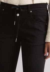 Person in schwarzen Hosen mit einer Kordelzug-Taille, die zu einer Schleife gebunden ist, kombiniert mit einem cremefarbenen Strickpullover, Hand entspannt an der Seite.