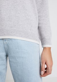 Grauer Strickpullover mit einem weißen unteren Saum und Bündchen, gezeigt über einer hellblauen Jeans. Nahaufnahme von Ärmeln und Handdetails.