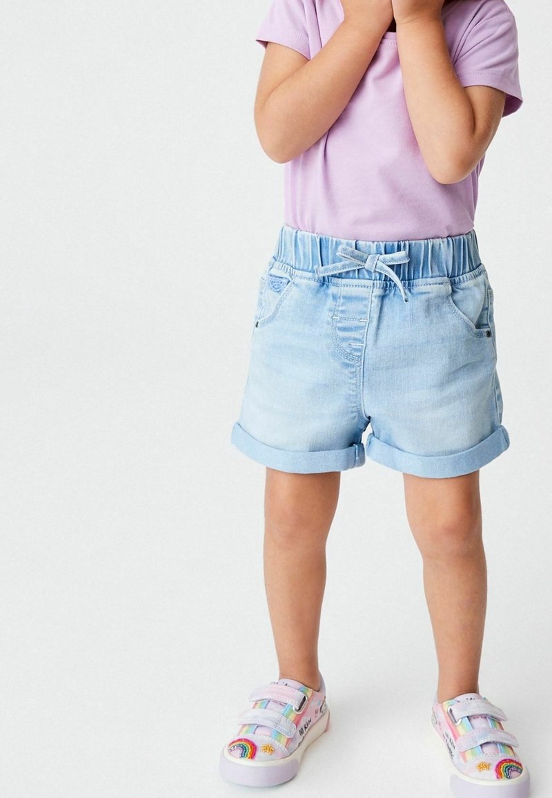 Enfant portant un t-shirt violet clair, un short en jean bleu clair roulé et des baskets colorées avec des arcs-en-ciel, debout avec les mains près du visage.