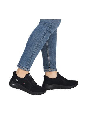 Person trägt schwarze Sportsneaker und blaue Jeans und steht mit einem leicht gehobenen Fuß.