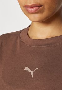 Pruun puuvillane T-särk, millel on ümar kaelus ja vasakul rinnal on heled värviga hüppav kassi logo. Pehme tekstuur.