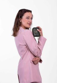Černá peněženka na zip s kulatým logem, držena osobou v jemně růžovém přizpůsobeném saku, stojící v profilu na bílém pozadí.