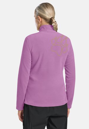 Vrouw met blond gevlochten haar die een paarse fleecejas draagt met een gele pootafdruk op de bovenrug, gecombineerd met een zwarte broek.